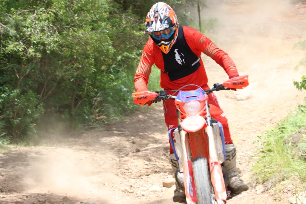 Motocross rider in red gear riding on a dirt trail with a forest background.