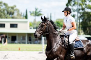 Equestrian rider in white attire and helmet riding a dark horse with focused expression during training.