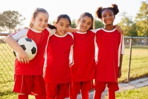 Four young girls in red soccer uniforms smiling with one holding a soccer ball