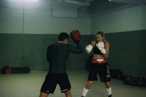A female boxer training with a male coach in a dimly lit gym, both wearing boxing gloves and focused on the exercise