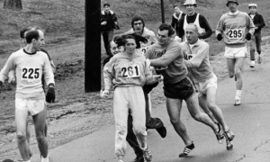 Black and white photo of marathon runners with one female athlete wearing number 261 being physically restrained by a race official