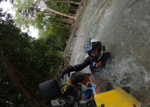 Adventurous motorcyclist in full body armor navigating a fast-moving river in a forested area.